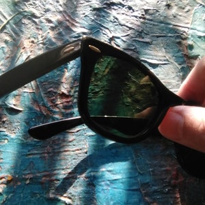 K&ouml;nnte beinhalten: Schwarze Sonnenbrille mit dunkel get&ouml;nten Gl&auml;sern und einem klassischen Rahmendesign. Die B&uuml;gel sind mit kleinen, metallisch silberfarbenen Akzenten an den Scharnieren mit dem Vorderrahmen verbunden, was dem dunklen Design ein dezentes Detail verleiht.
