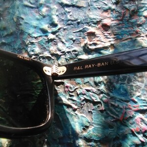 K&ouml;nnte beinhalten: Schwarze Sonnenbrille mit einem goldfarbenen Metallscharnier. Der innere B&uuml;gel tr&auml;gt die Aufschrift B&L RAY-BAN U.S. und der vordere Rahmen zeigt den Text B&L 5024. Das Design umfasst ein dunkles Glas und eine gl&auml;nzend schwarze Rahmenkonstruktion.