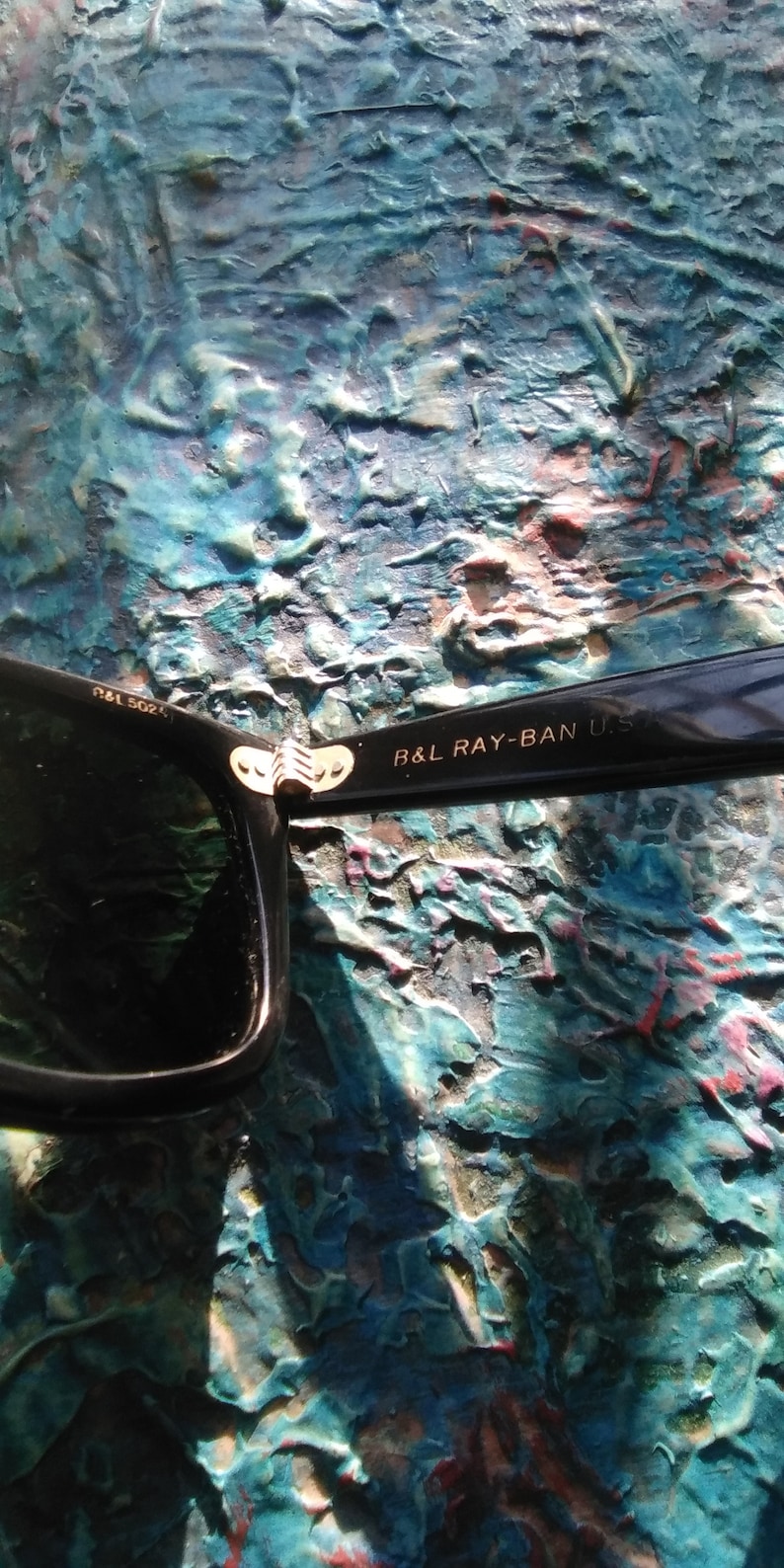 K&ouml;nnte beinhalten: Schwarze Sonnenbrille mit einem goldfarbenen Metallscharnier. Der innere B&uuml;gel tr&auml;gt die Aufschrift B&L RAY-BAN U.S. und der vordere Rahmen zeigt den Text B&L 5024. Das Design umfasst ein dunkles Glas und eine gl&auml;nzend schwarze Rahmenkonstruktion.