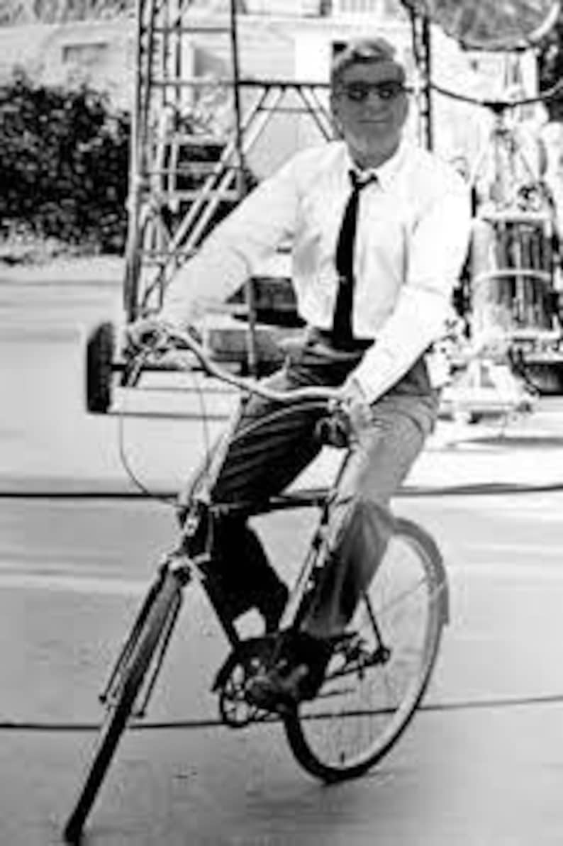 K&ouml;nnte beinhalten: Ein Schwarz-Wei&szlig;-Foto einer Person, die ein helles, lang&auml;rmeliges Hemd, eine dunkle Krawatte und eine dunkle Hose tr&auml;gt und auf einem Fahrrad sitzt. Die Person tr&auml;gt eine Brille mit dunklem Rahmen und schaut in die Kamera.