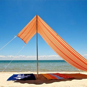 K&ouml;nnte beinhalten: Ein orange-wei&szlig; gestreiftes Strand-Sonnensegel, das von einem zentralen Mast mit wei&szlig;en Abspannleinen gehalten wird. Unter dem Segel liegen ein dunkelblaues Handtuch mit wei&szlig;em Grafikdesign und ein mehrfarbig gestreiftes Handtuch flach im Sand.