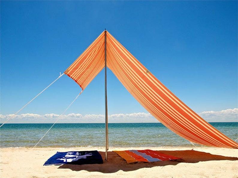 K&ouml;nnte beinhalten: Ein orange-wei&szlig; gestreiftes Strand-Sonnensegel, das von einem zentralen Mast mit wei&szlig;en Abspannleinen gehalten wird. Unter dem Segel liegen ein dunkelblaues Handtuch mit wei&szlig;em Grafikdesign und ein mehrfarbig gestreiftes Handtuch flach im Sand.