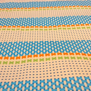 May include: A white fabric with a woven pattern of blue, orange, and green stripes. The fabric has a subtle texture and a slightly wavy appearance.