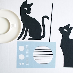 K&ouml;nnte beinhalten: Eine wei&szlig;e Porzellansaucers mit einer wei&szlig;en Porzellan Tasse gef&uuml;llt mit einer wei&szlig;en Fl&uuml;ssigkeit. Zwei schwarze Silhouette Katzen sind auf einem wei&szlig;en Hintergrund mit einem blauen und wei&szlig;en Radio in der Mitte.