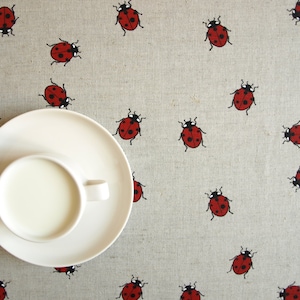 May include: A white teacup and saucer on a beige tablecloth decorated with red ladybugs. The cup contains a white liquid, perhaps milk or cream. The ladybugs feature black spots and details.