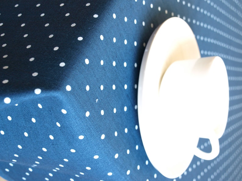 May include: A white teacup and saucer on a blue tablecloth with white polka dots.