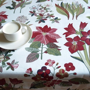 May include: A white ceramic teacup and saucer with a white floral pattern tablecloth. The tablecloth has red and green floral designs.