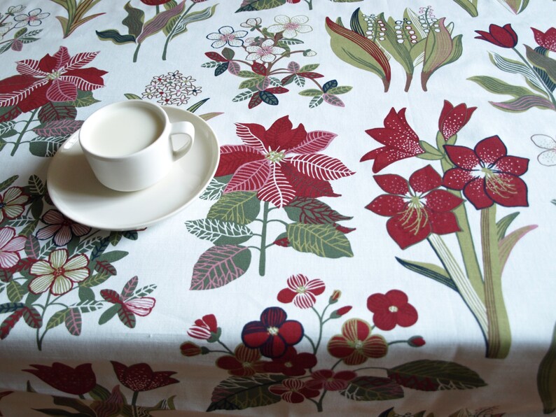 May include: A white ceramic teacup and saucer with a white floral pattern tablecloth. The tablecloth has red and green floral designs.