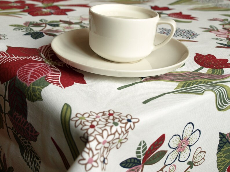 May include: A white ceramic coffee cup with a white saucer, sitting on a white tablecloth with a floral pattern in shades of red, green, and white.