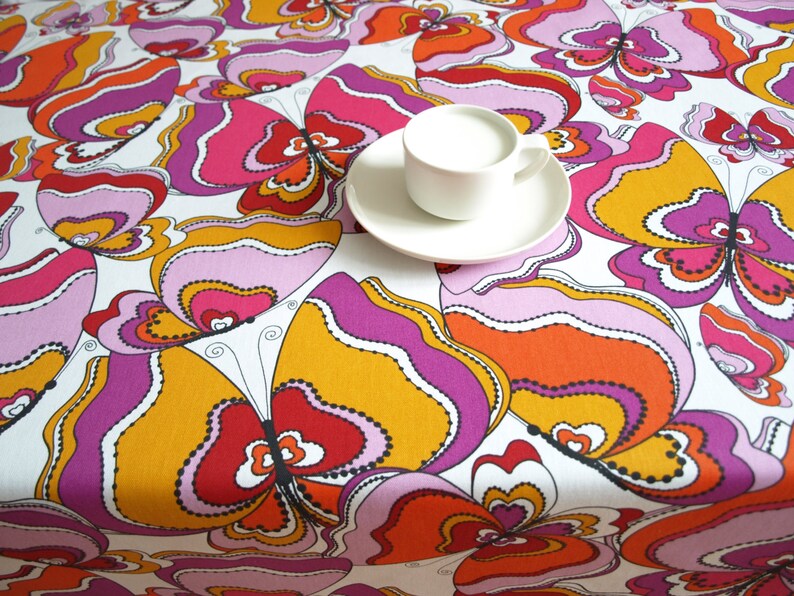May include: A white tablecloth with a colorful butterfly pattern in shades of pink, orange, and yellow. The tablecloth features a stylized butterfly design with intricate details.