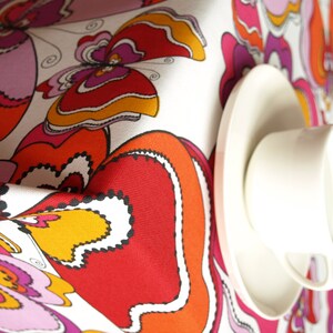 May include: White tablecloth with a colorful butterfly pattern in shades of red, orange, pink, and yellow. A white teacup and saucer are placed on the tablecloth.