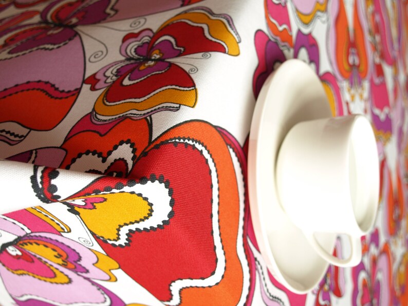 May include: White tablecloth with a colorful butterfly pattern in shades of red, orange, pink, and yellow. A white teacup and saucer are placed on the tablecloth.