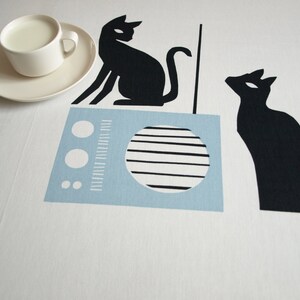 K&ouml;nnte beinhalten: Eine wei&szlig;e Tischdecke mit einem blau-wei&szlig;en Radio, zwei schwarzen Katzen-Silhouetten und einer wei&szlig;en Untertasse mit einer wei&szlig;en Tasse gef&uuml;llt mit Milch.