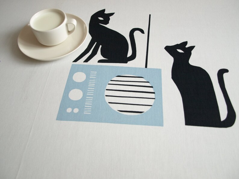K&ouml;nnte beinhalten: Eine wei&szlig;e Tischdecke mit einem blau-wei&szlig;en Radio, zwei schwarzen Katzen-Silhouetten und einer wei&szlig;en Untertasse mit einer wei&szlig;en Tasse gef&uuml;llt mit Milch.