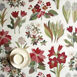 May include: A white tablecloth with a floral pattern featuring red, green, and white flowers. A white saucer with a white cup sits on the tablecloth.