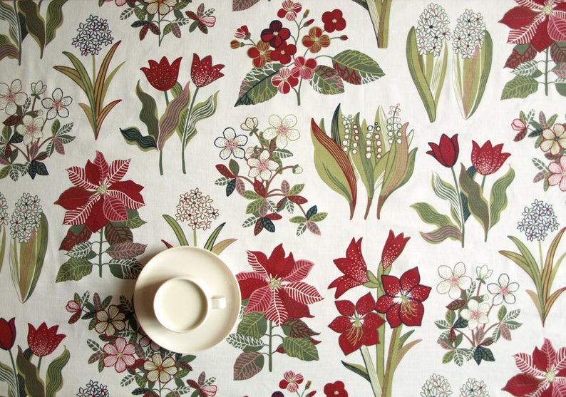 May include: A white tablecloth with a floral pattern featuring red, green, and white flowers. A white saucer with a white cup sits on the tablecloth.