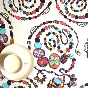 May include: A white tablecloth with a colourful pattern of beads and circles. The pattern is reminiscent of a necklace or bracelet. A white saucer with a white cup sits on the tablecloth.