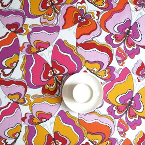 May include: A white tablecloth with a repeating pattern of colorful butterflies. The butterflies are pink, orange, red, and yellow with black outlines. A white saucer is in the center of the tablecloth.