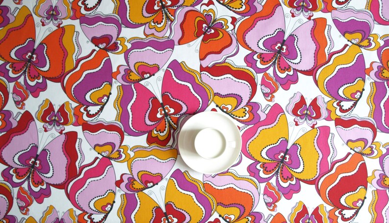 May include: A white tablecloth with a repeating pattern of colorful butterflies. The butterflies are pink, orange, red, and yellow with black outlines. A white saucer is in the center of the tablecloth.