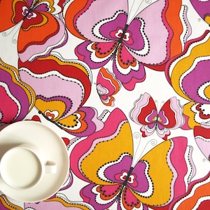 May include: A white tablecloth with a repeating pattern of colorful butterflies. The butterflies are pink, orange, yellow, and red with black outlines. The tablecloth is perfect for a picnic or a party.