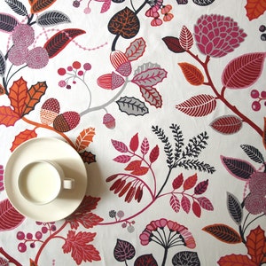 May include: A white tablecloth with a floral pattern in shades of red, pink, black, and gray. The pattern features leaves, berries, and flowers. A white saucer with a white cup sits on the tablecloth.