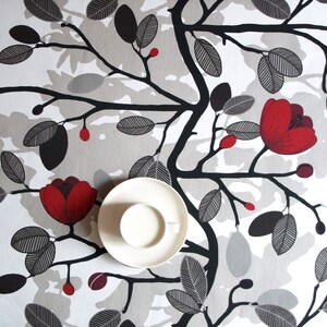 May include: A white tablecloth with a black and gray floral pattern. The flowers are red and the leaves are gray. A white saucer with a white cup sits on the tablecloth.