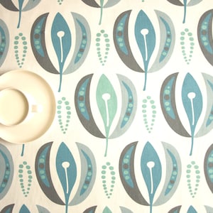 May include: A white ceramic saucer with a white ceramic cup filled with a light-colored liquid. The saucer and cup are on a white tablecloth with a repeating pattern of blue, gray, and green stylized leaves.