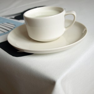 K&ouml;nnte beinhalten: Eine wei&szlig;e Keramiktasse mit Untertasse, gef&uuml;llt mit einer wei&szlig;en Fl&uuml;ssigkeit, steht auf einer wei&szlig;en Tischdecke mit einem schwarzen und blauen geometrischen Muster.