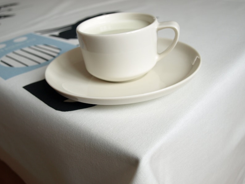 K&ouml;nnte beinhalten: Eine wei&szlig;e Keramiktasse mit Untertasse, gef&uuml;llt mit einer wei&szlig;en Fl&uuml;ssigkeit, steht auf einer wei&szlig;en Tischdecke mit einem schwarzen und blauen geometrischen Muster.
