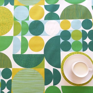 Peut inclure: Une nappe blanche avec un motif géométrique de cercles et de demi-cercles verts, jaunes et bleus. Une soucoupe blanche avec une tasse blanche est posée sur la nappe.