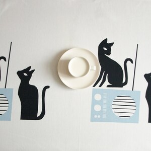 K&ouml;nnte beinhalten: Eine wei&szlig;e Tischdecke mit vier schwarzen Katzensilhouetten. Zwei der Katzen sitzen auf blauen und wei&szlig;en Radios. Eine wei&szlig;e Untertasse mit einer Tasse Milch befindet sich in der Mitte der Tischdecke.