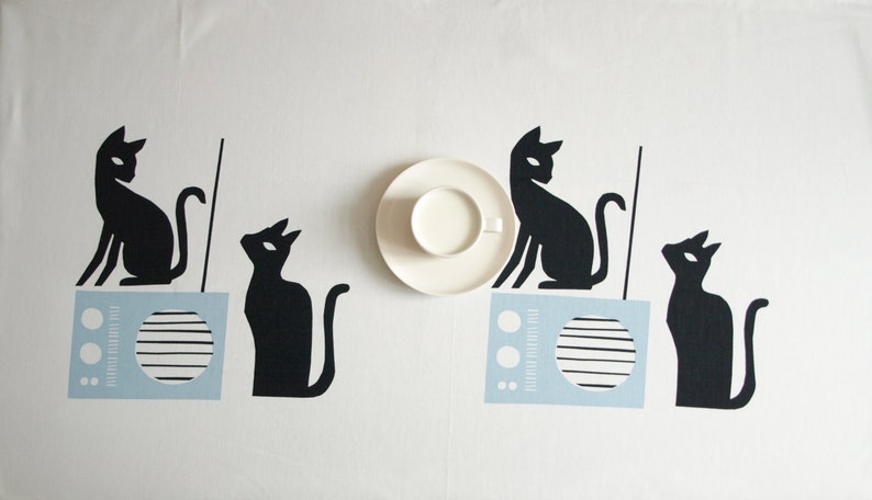 K&ouml;nnte beinhalten: Eine wei&szlig;e Tischdecke mit vier schwarzen Katzensilhouetten. Zwei der Katzen sitzen auf blauen und wei&szlig;en Radios. Eine wei&szlig;e Untertasse mit einer Tasse Milch befindet sich in der Mitte der Tischdecke.