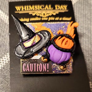 May include: A rectangular pin featuring a black witch hat and a purple cauldron with orange flames. The pin has a background with purple and orange paint splatters and the text CAUTION! in pink. It is mounted on a black display card with the text WHIMSICAL DAY and the shop URL.