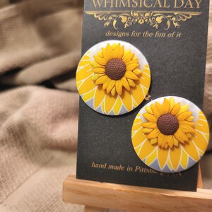 May include: Two round sunflower earrings with yellow petals and brown centers are displayed on a black card. The card reads "WHIMSICAL DAY" and "designs for the fun of it." The text "hand made in Pittsburgh" is also visible.
