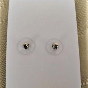 May include: Two silver-colored stud earrings with clear plastic backings are displayed on a white card. The earrings have a small, round design. The background is a neutral color.