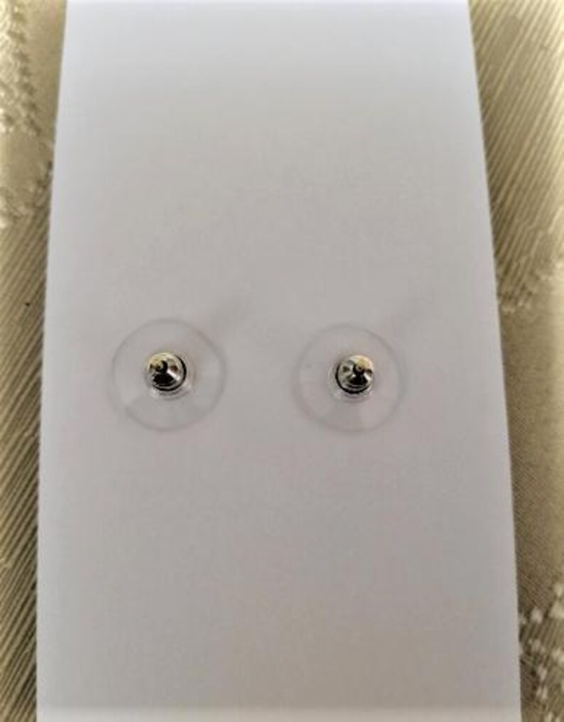 May include: Two silver-colored stud earrings with clear plastic backings are displayed on a white card. The earrings have a small, round design. The background is a neutral color.