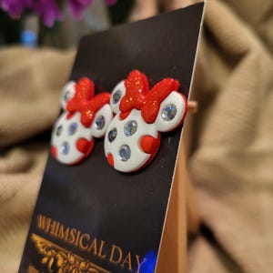 May include: A pair of whimsical stud earrings featuring a white and red design with sparkling accents. The earrings are shaped like a cartoon character with a red bow and polka dots. The earrings are displayed on a black card that reads "Whimsical Day".