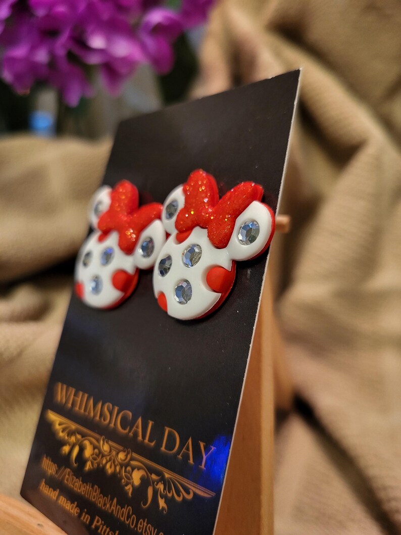 May include: A pair of whimsical stud earrings featuring a white and red design with sparkling accents. The earrings are shaped like a cartoon character with a red bow and polka dots. The earrings are displayed on a black card that reads "Whimsical Day".