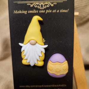 May include: A whimsical pin set featuring a yellow gnome with a white beard and a purple Easter egg with yellow floral accents. The text "WHIMSICAL DAY" and "Making smiles one pin at a time!" are displayed on the black backing.