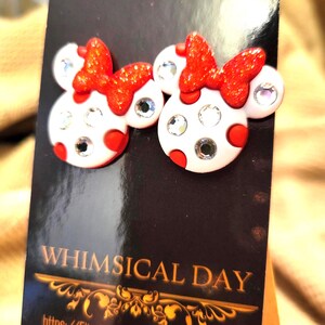 May include: A pair of whimsical stud earrings featuring a Minnie Mouse design. The earrings have a white base with red polka dots, sparkling red bows, and clear crystal accents. The earrings are displayed on a black card with the text "WHIMSICAL DAY".