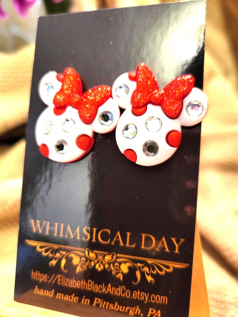 May include: A pair of whimsical stud earrings featuring a Minnie Mouse design. The earrings have a white base with red polka dots, sparkling red bows, and clear crystal accents. The earrings are displayed on a black card with the text "WHIMSICAL DAY".