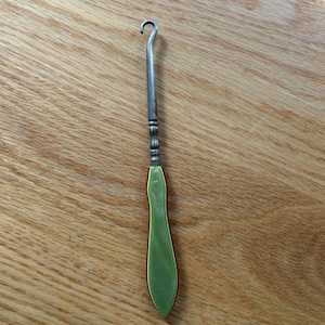 May include: A button hook featuring a metallic silver-toned shaft with a curved tip and a smooth, light green handle. The handle has a tapered, elongated shape with a polished finish, connecting to the metal hook via a decorative, ridged collar.