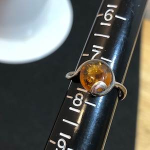 May include: A silver ring featuring a round, translucent amber stone set on a black ring mandrel. The metal band displays a swirling design on one side of the stone. The ring is positioned at the 8-inch mark on the black measuring tool.