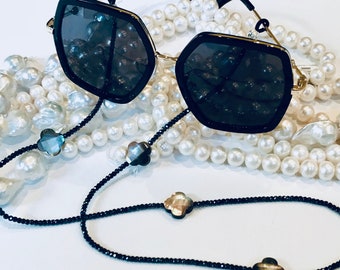 May include: Black sunglasses with a gold frame and black beaded chain with iridescent flower accents. The sunglasses are resting on a strand of white pearls.