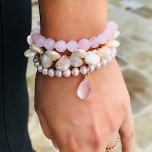 Könnte beinhalten: Ein Set aus drei Armbändern mit rosa und weißen Perlen. Das obere Armband besteht aus rosa Rosenquarzperlen, das mittlere Armband aus weißen Perlen und das untere Armband aus rosa Perlen. Ein rosa Rosenquarz-Anhänger hängt am unteren Armband.