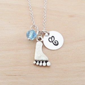 May include: Silver necklace with a charm of a footprint, a blue crystal bead, and a silver disc with the letter 'L' on it.