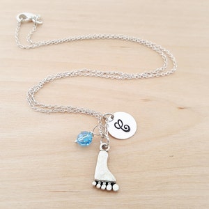 May include: A silver necklace with a charm of a footprint, a small blue bead, and a silver disc with the letter 'E' on it.