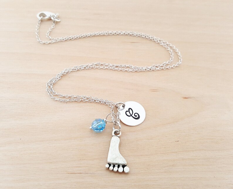 May include: A silver necklace with a charm of a footprint, a small blue bead, and a silver disc with the letter 'E' on it.