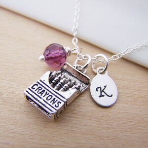 May include: Silver necklace with a charm of a box of crayons, a purple crystal bead, and a silver charm with the letter K.