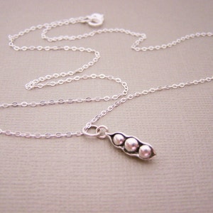 May include: A delicate silver necklace featuring a pea pod pendant with three small, round peas. The chain is thin and the pendant is detailed. The necklace is displayed on a neutral background.
